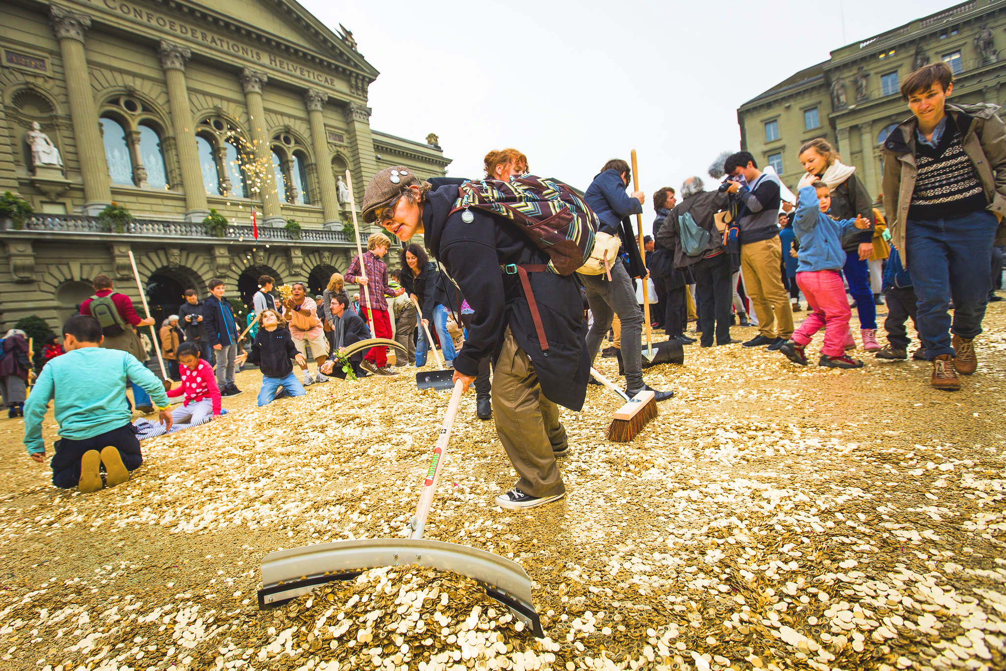 Une foule de gens sont réunis sur une place. Il y a des pièces de monnaie partout sur le sol, certains jouent avec, d'autres balaient.