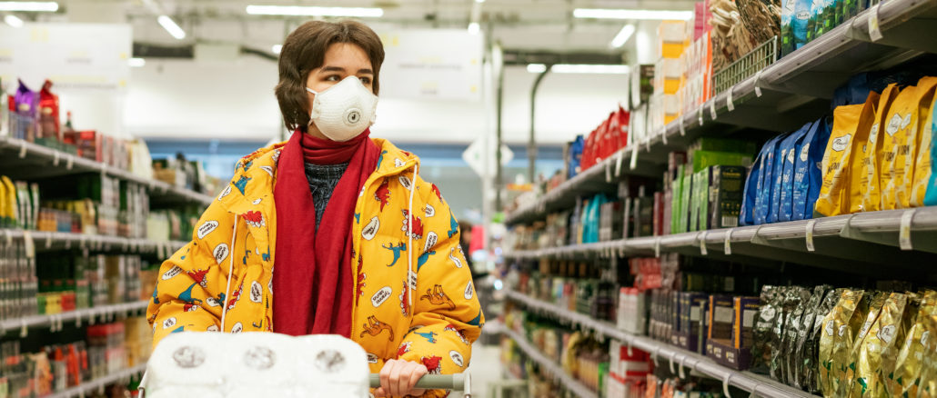 Un jeune femme est en train de faire ses courses dans un supermarché. Elle est masquée et son caddie est remplie de papier toilette.