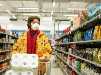 Un jeune femme est en train de faire ses courses dans un supermarché. Elle est masquée et son caddie est remplie de papier toilette.